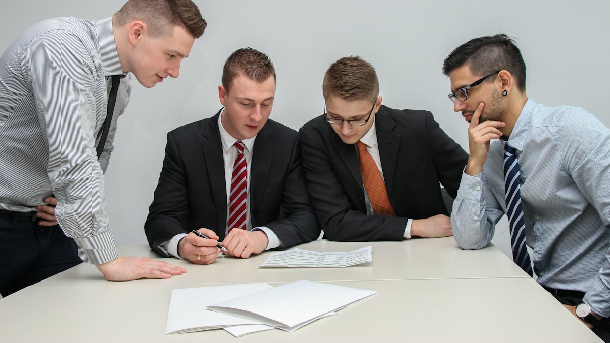 Quatro homens de camisa e gravata reunidos à mesa observando papéis