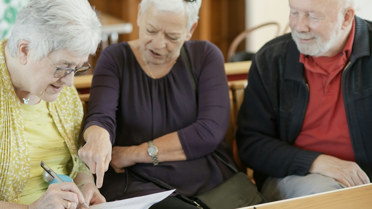 Três pessoas idosas sentadas lado a lado analisando juntas um papel sobre a mesa