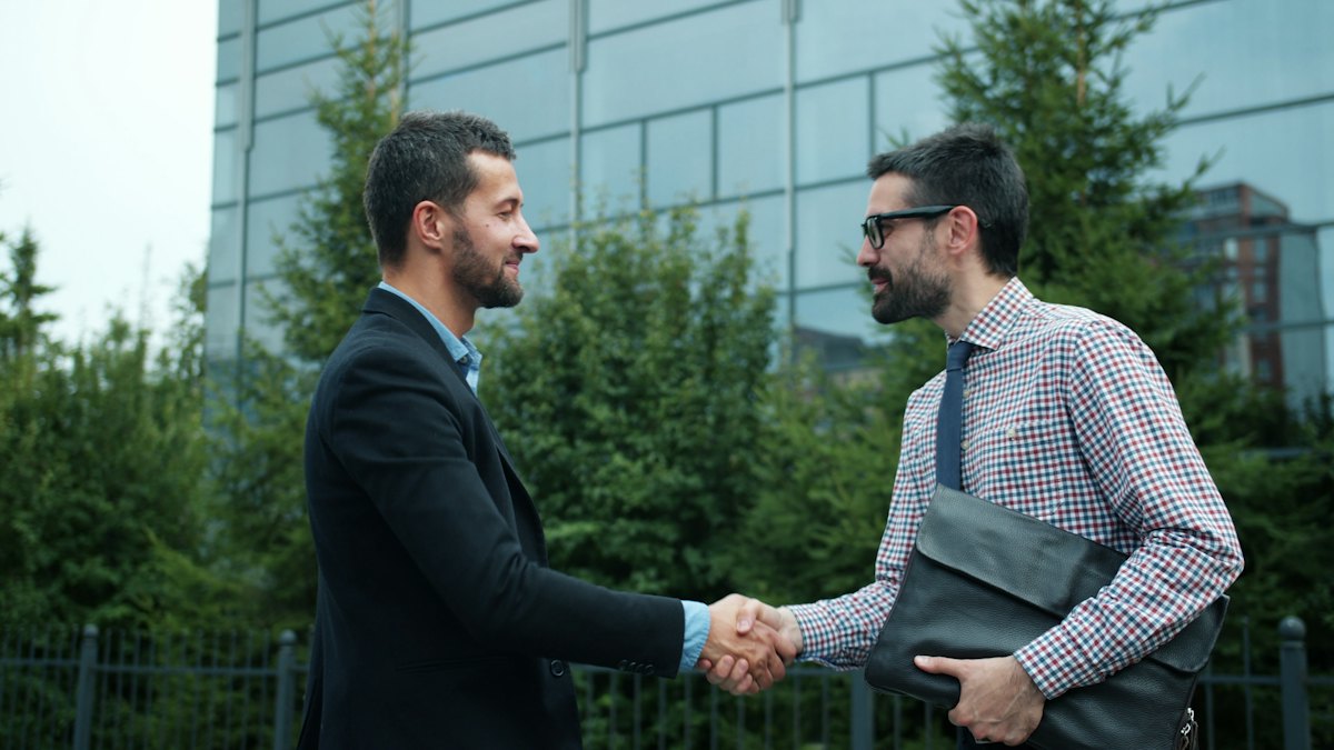 Dois homens de roupa social apertando as mãos ao ar livre diante de árvores