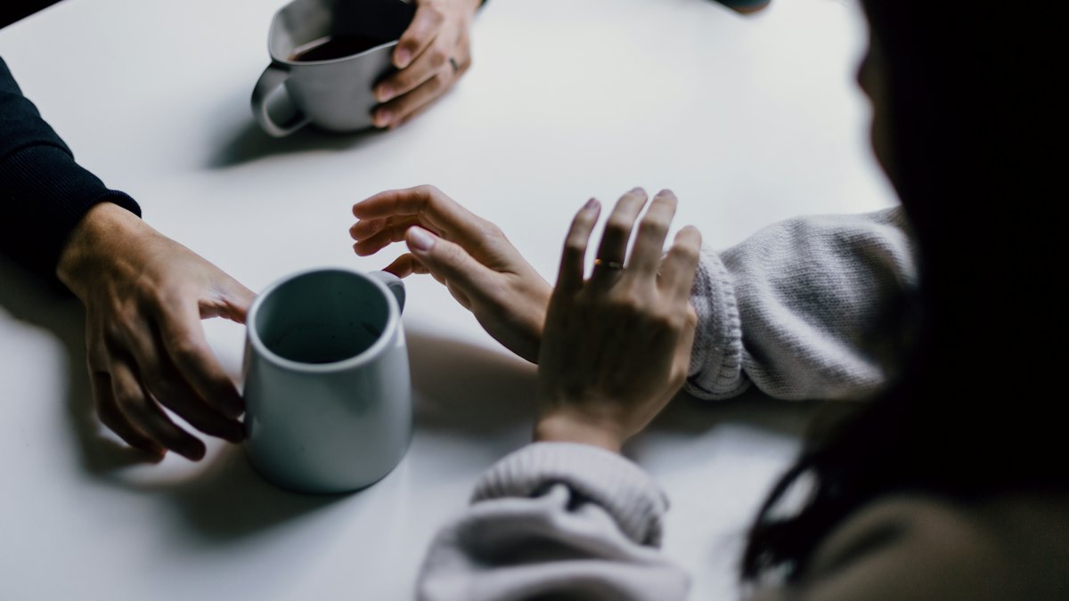 Mãos de pessoas ao redor de uma mesa branca segurando canecas de café durante conversa
