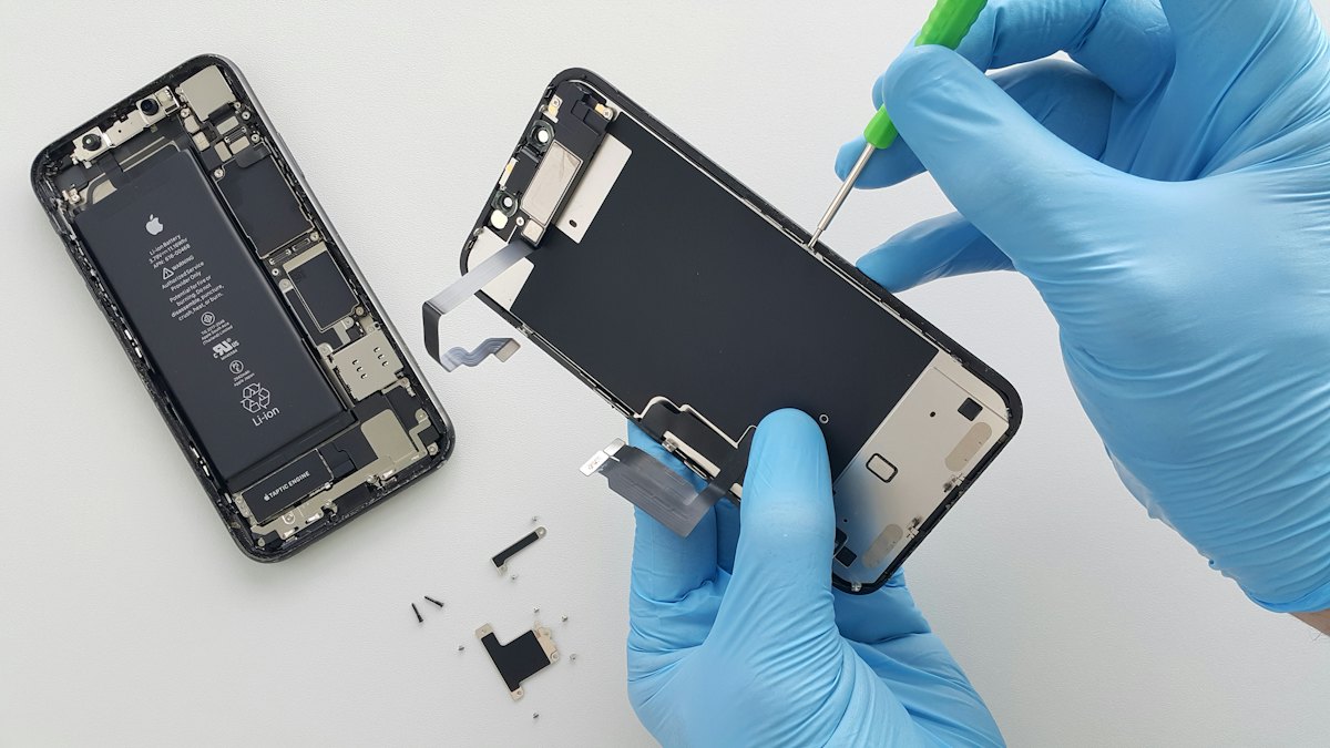 Mãos com luvas azuis consertando um celular aberto com peças internas à mostra