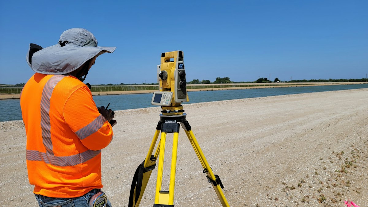 Topógrafo de colete laranja ao lado de um equipamento de medição sobre tripé amarelo