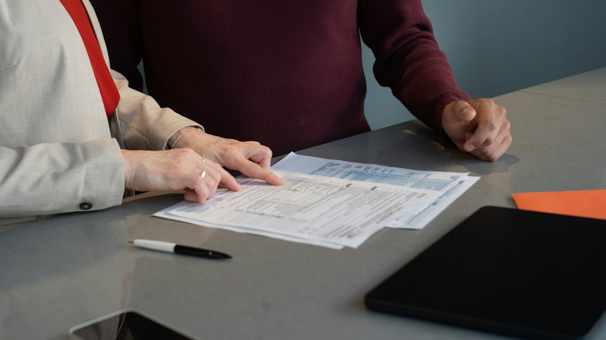 Duas pessoas sentadas a uma mesa cinza revisando juntas documentos impressos